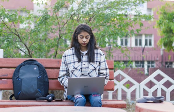 Brunette young student in white and black flannel on laptop outside on a brown bench