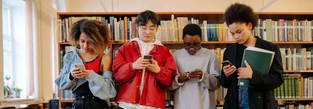 Four students all looking down at their phones in front of library books.
