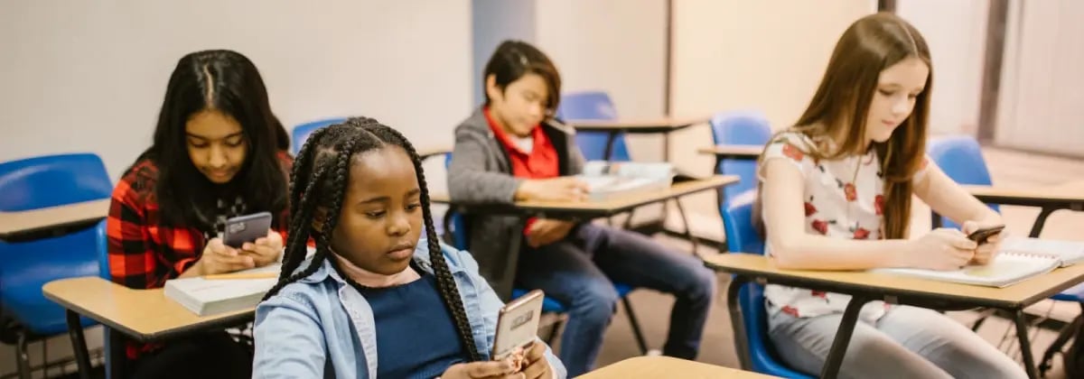Four young learners all preoccupied on phones sitting at desks in the classroom.