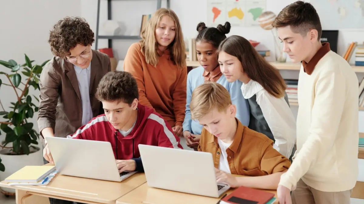 Class of students with two sitting behind laptops and peers standing behind them collaborating on a project.