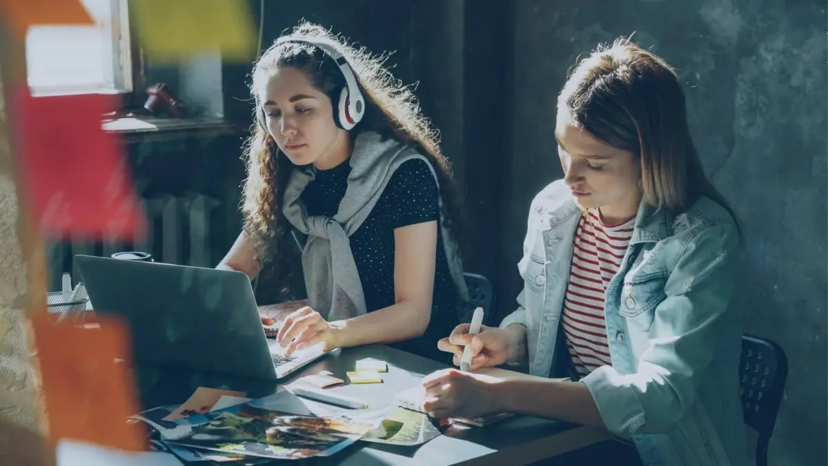 Two female students sitting next to each other: One with headphones and writing on a laptop and the other storyboarding next to her.