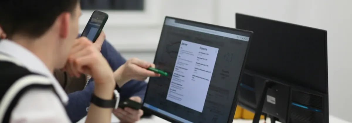 Young male looking at computer screen with someone pointing next to him holding a mobile device.