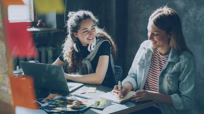 Two female learners smiling while working alongside each other by an open laptop.