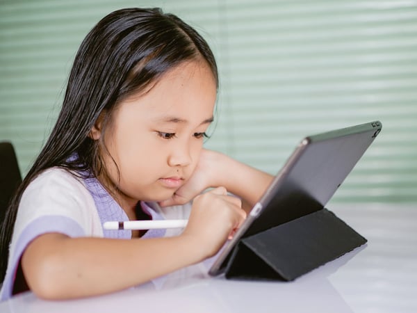 Young K-5 student sitting at a tablet completing an assignment with tool in hand.