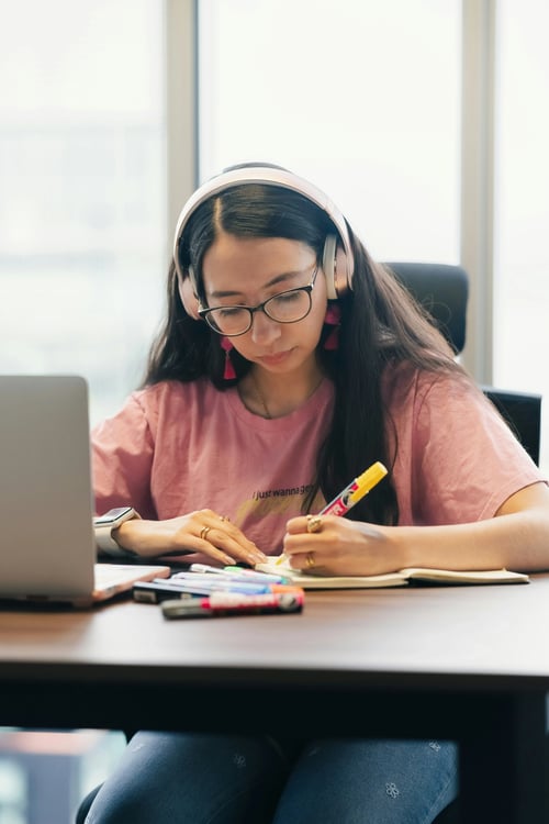 Female learner with pink shirt, pink earrings, pink headphones, and black glasses highlighting something in their journal with their laptop open.