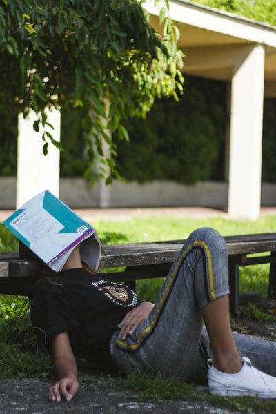 Learner with book over their face while laying down and resting on a bench outside.